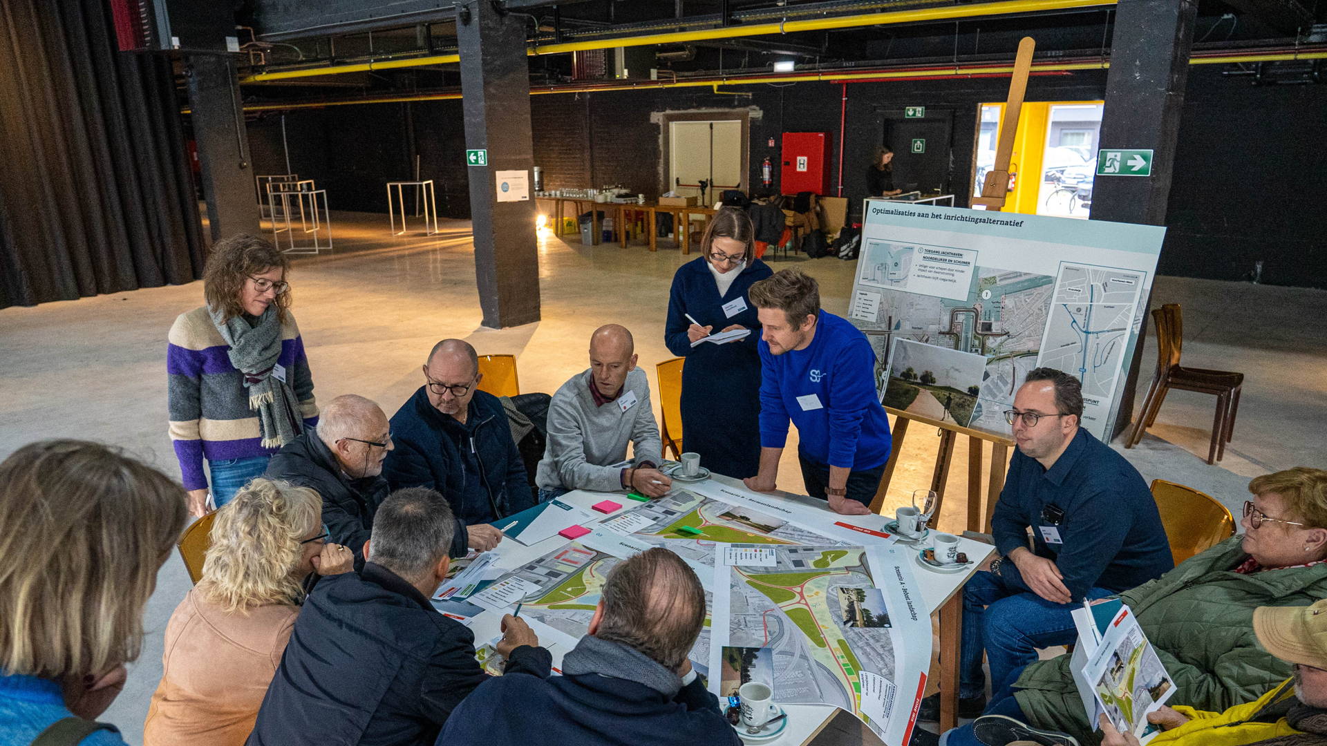 Une dizaine de personnes est assise autour d'une table sur laquelle sont posés plusieurs plans lors d'une séance d'information du projet Nieuwe Sluis Zeebrugge.