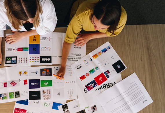 Twee vrouwen zitten aan een tafel met papieren met logo's erop