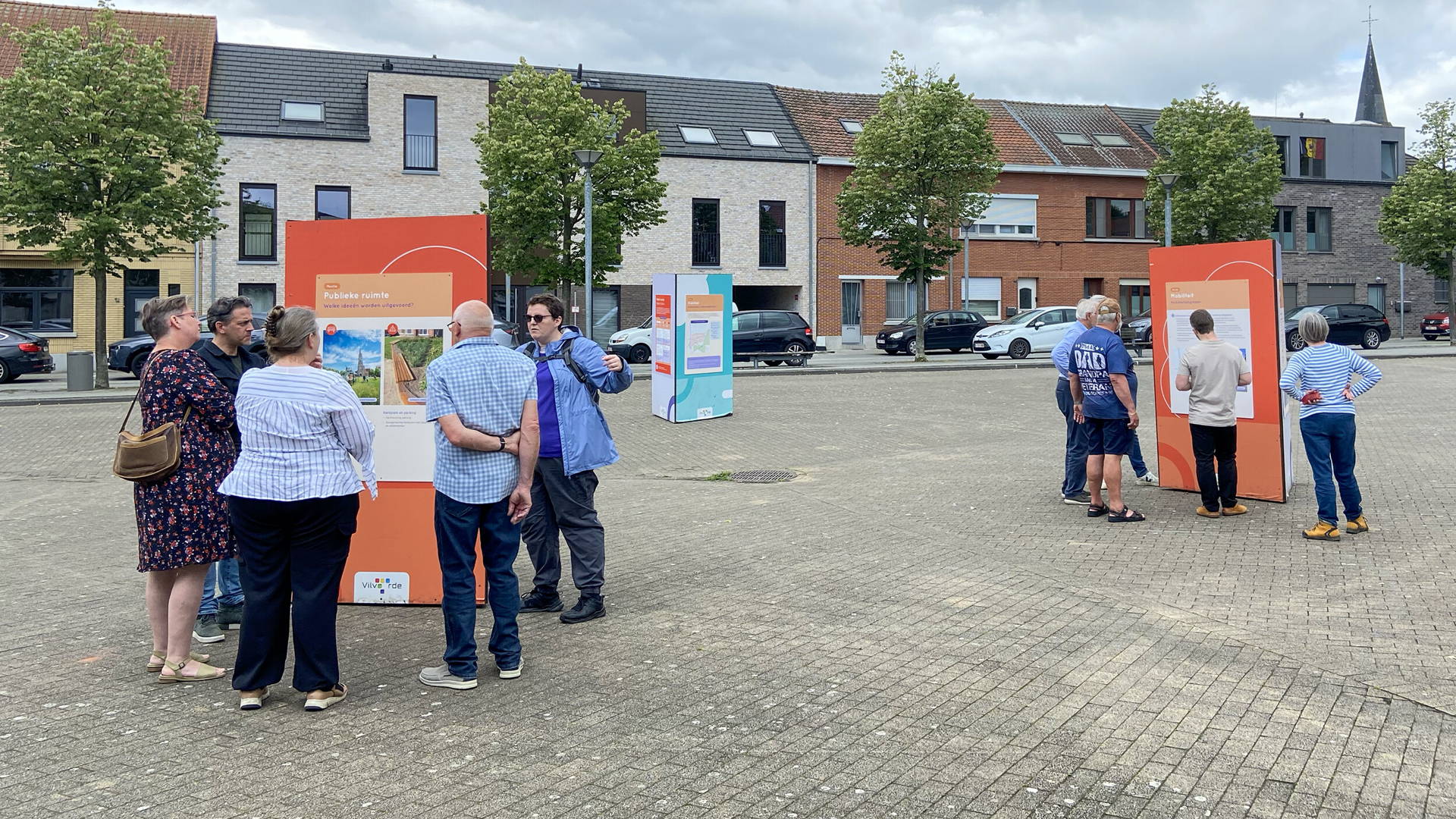 Informatieborden staan verspreid op een plein in Vilvoorde Peutie. De informatieborden staan in een driehoek en de mensen staan er rond. De mensen lezen de informatie op de informatieborden.
