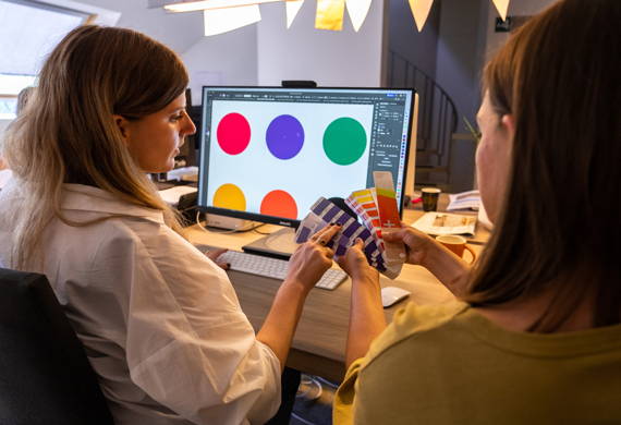 Twee vrouwen zitten voor computerscherm en bespreken verschillende kleurstalen