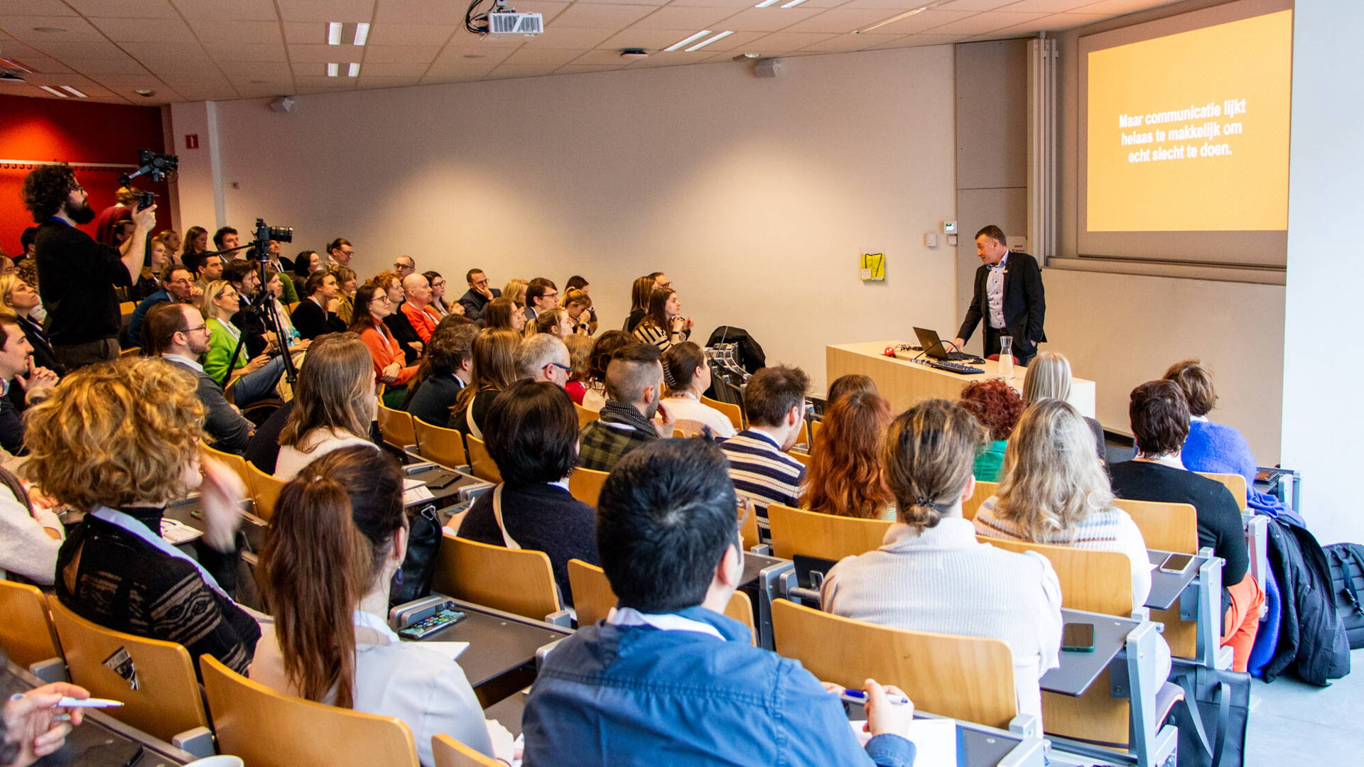 Kortom studiedag omgevingscommunicatie in de hogeschool inovant Antwerpen. Een aula zit vol met personen en luisteren naar de presentator vooraan de aula.