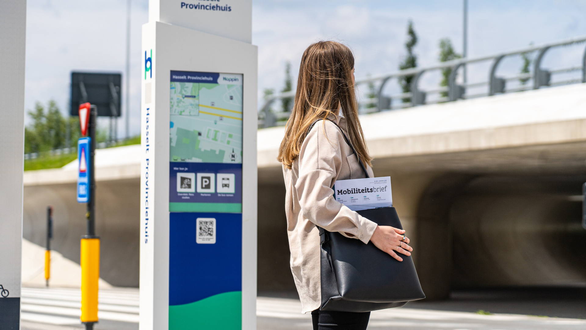 Vrouw heeft de mobiliteitsbrief vast in haar rechterhand. Loopt over straat met op de achtergrond een hoppinpunt een brug.